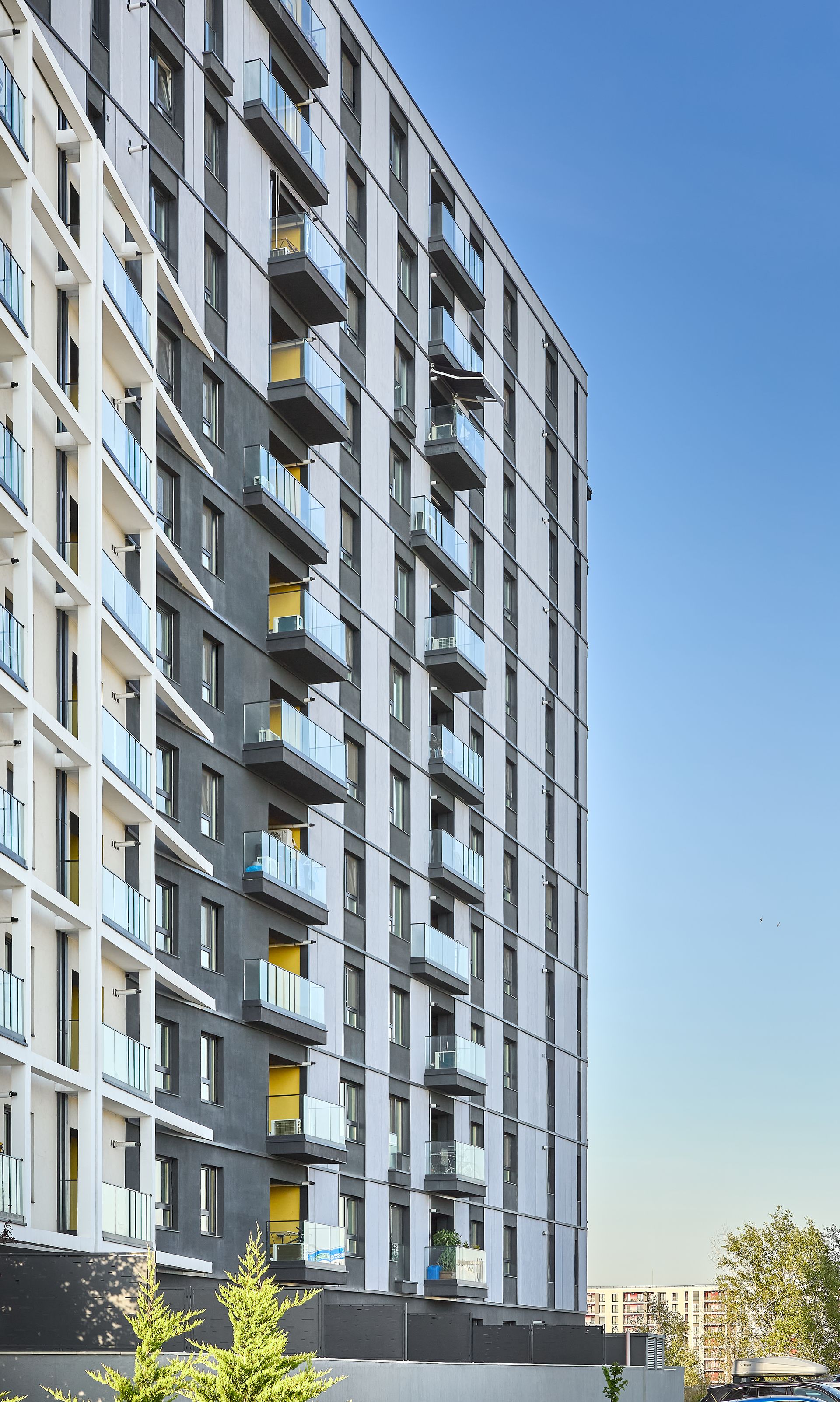 Vedere verticală în perspectivă de-a lungul fațadei placate cu panouri gri-antracit, surprinsă de la nivelul solului privind în sus. Se remarcă ritmul balcoanelor în consolă cu balustrade din sticlă, panourile de fibrociment cu accente de tencuială albă, nișele balcoanelor în tonuri galbene și efectul de perspectivă care accentuează înălțimea și registrele verticale ale ferestrelor.
