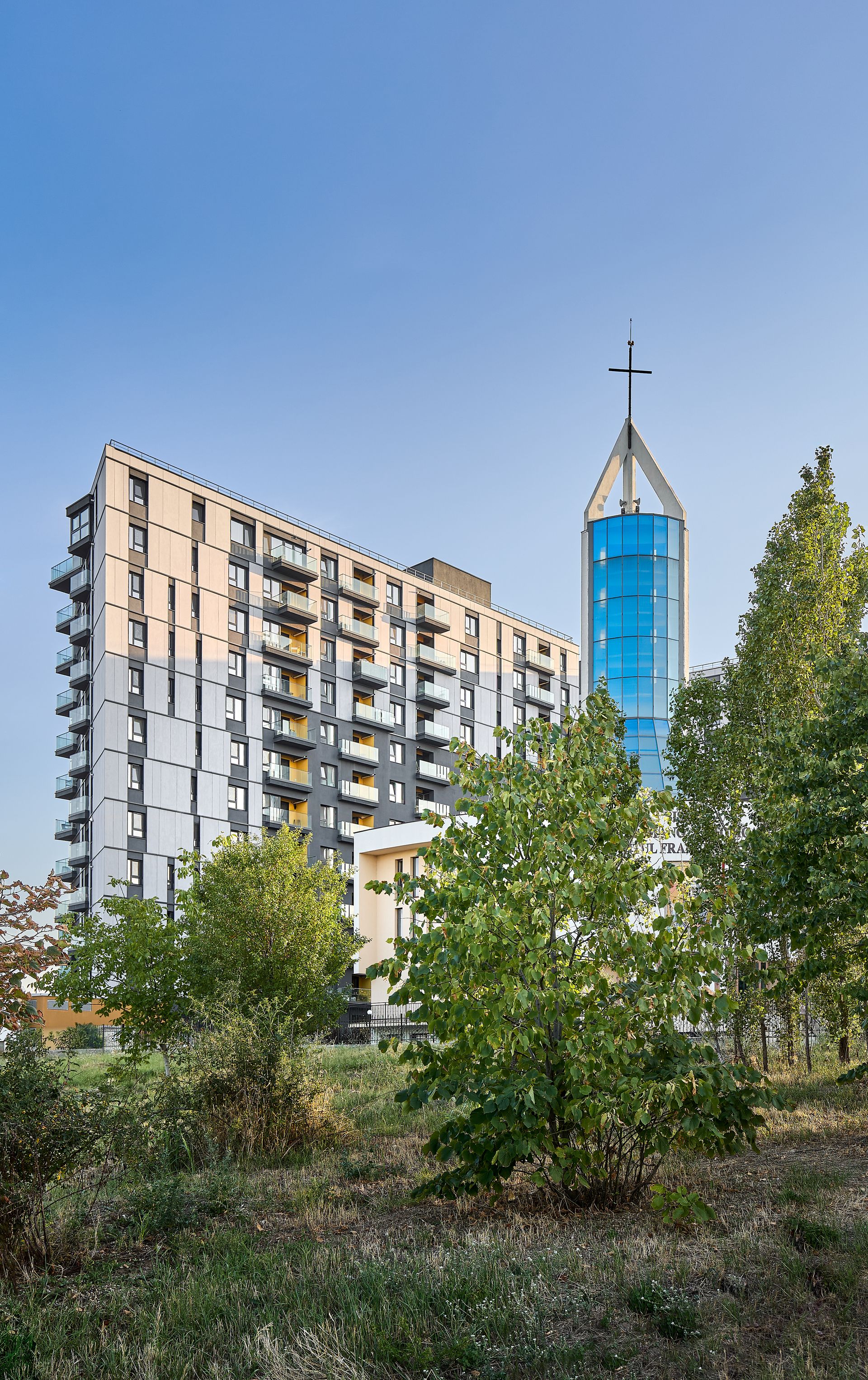 Imagine contextuală a clădirii, privită de la distanță peste o zonă verde amenajată, cu arbori și tufișuri în prim-plan. Alături de corpul rezidențial se ridică turnul din sticlă al bisericii moderne, cu flèche vitrată albastră terminată printr-o cruce, ilustrând integrarea ansamblului în cadrul natural și urban al vecinătății.