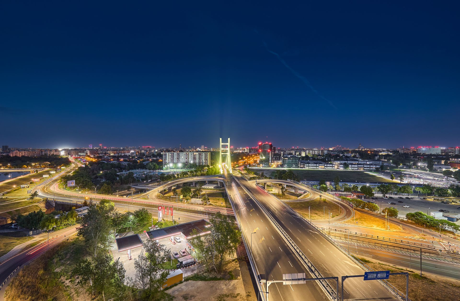 Panoramă nocturnă a zonei înconjurătoare: pilonul iluminat al podului hobanat domină priveliștea, în timp ce arterele și nodul rutier sunt marcate de dârele de lumină ale mașinilor. Se disting o benzinărie, spații verzi și silueta Bucureștiului până la orizont, sub un cer albastru închis — o imagine de context care evidențiază amplasarea strategică a ansamblului, lângă lac și marile artere de circulație.