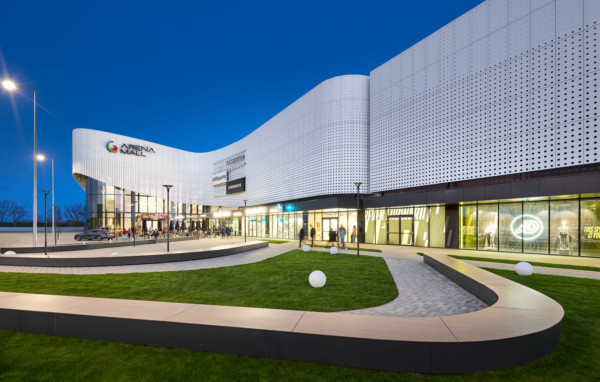 Fațada iluminată seara a Arena Mall Bacău, cu aleea de acces