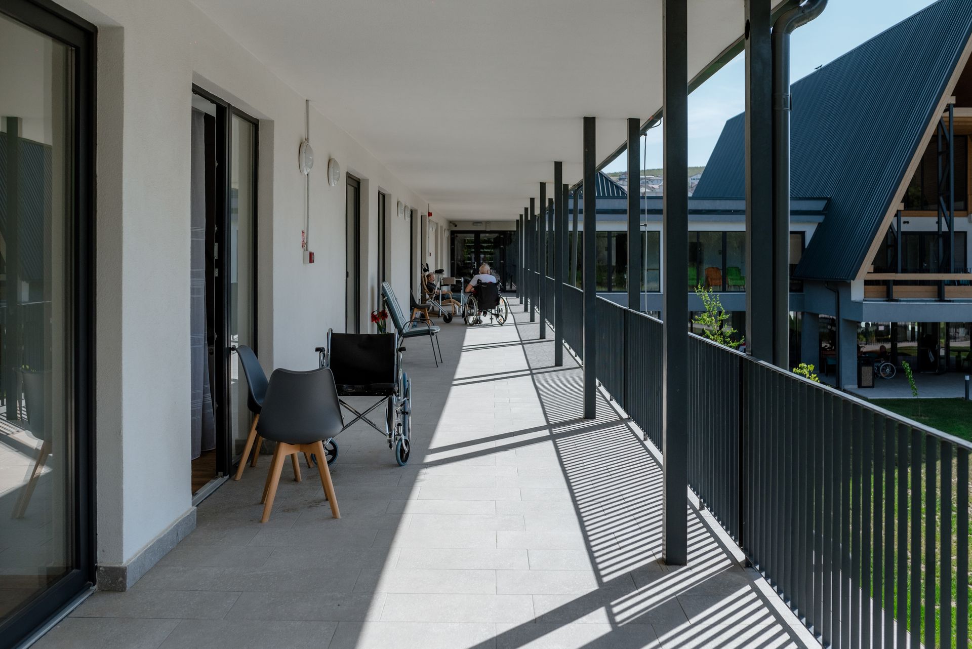 Coridorul-galerie exterior acoperit de la etaj, cu perete tencuit alb și uși glisante vitrate spre camere pe o latură, iar pe cealaltă stâlpi și balustradă metalică ce se deschid spre curte. Pardoseală din piatră; scaune și un scaun cu rotile marchează utilizarea de rezidență pentru seniori, cu aripa cu acoperișuri în pantă vizibilă peste curte.