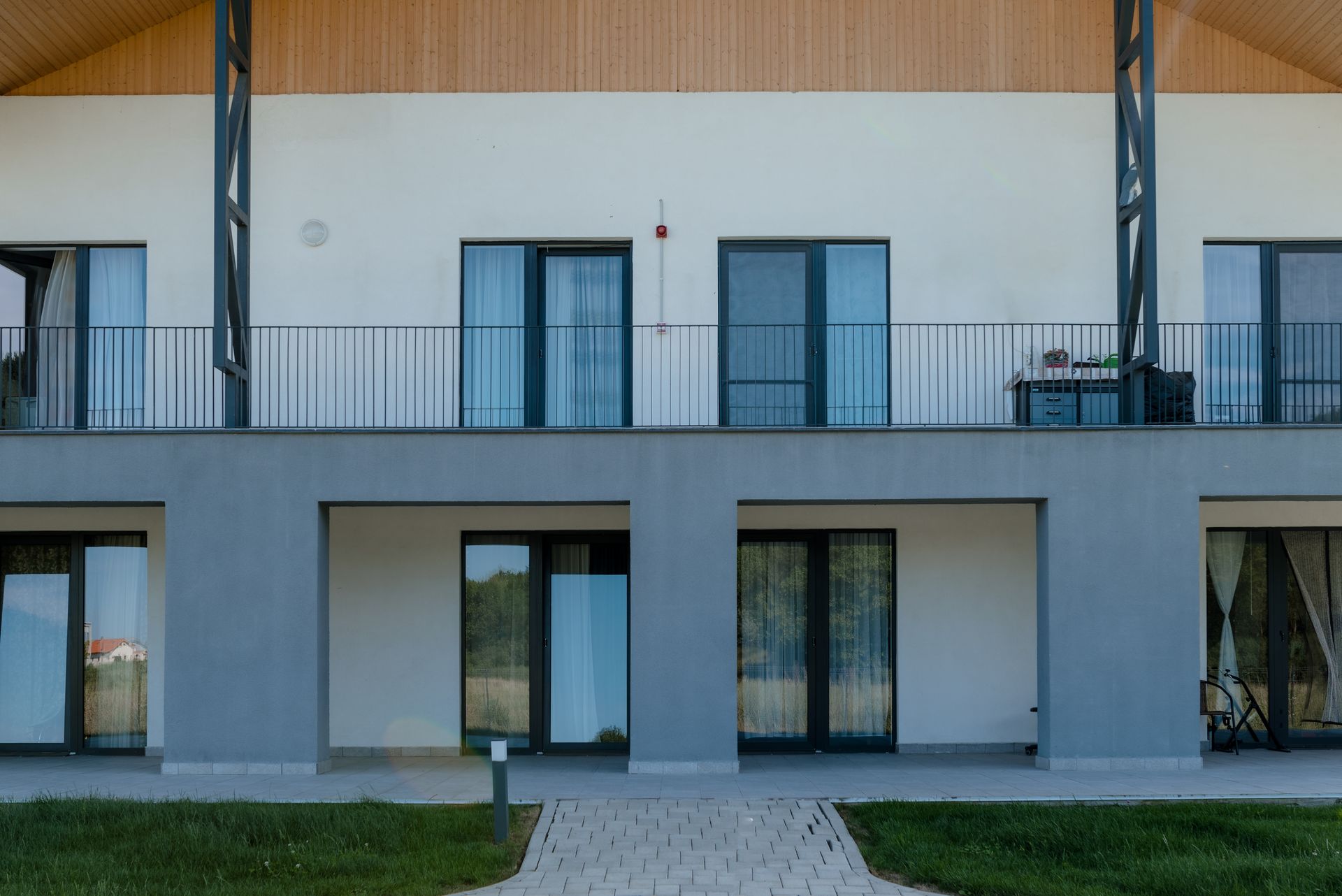Fațada rezidențială văzută frontal: la parter, loggii/terase retrase, tencuite gri, cu uși vitrate ample; la etaj, balcon continuu cu balustradă metalică zveltă și ferestre pe toată înălțimea. Deasupra, timpanul din lemn al acoperișului în pantă și contravântuirile metalice oblice; în prim-plan, peluză și alee pavată.