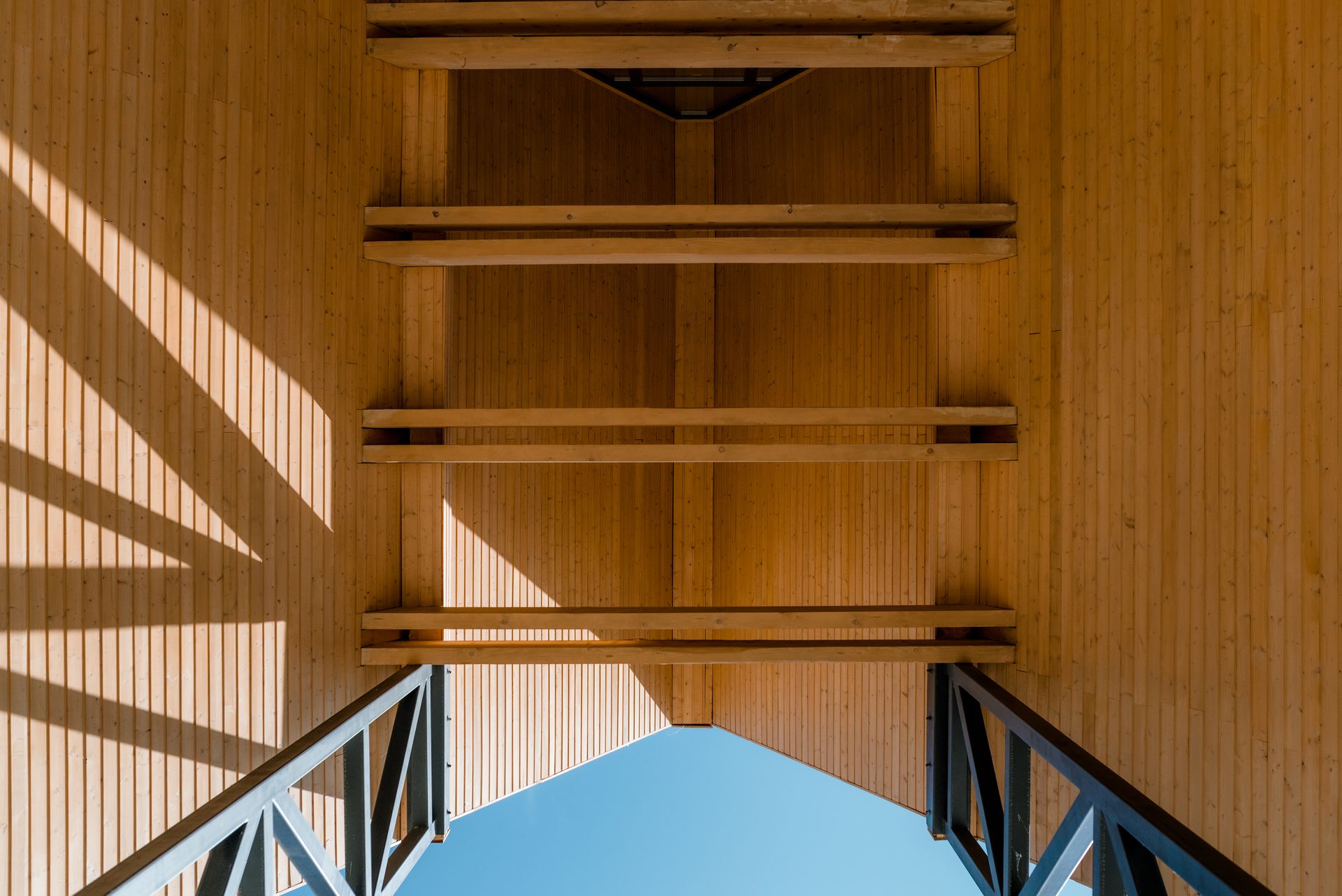 Detaliu arhitectural privit în sus către structura acoperișului: placarea din lemn natural pe verticală și grinzile orizontale din lemn, susținute de elemente metalice de contravântuire, cadrează o deschidere triunghiulară spre cerul albastru. Jocul de lumină și umbră accentuează textura lemnului.